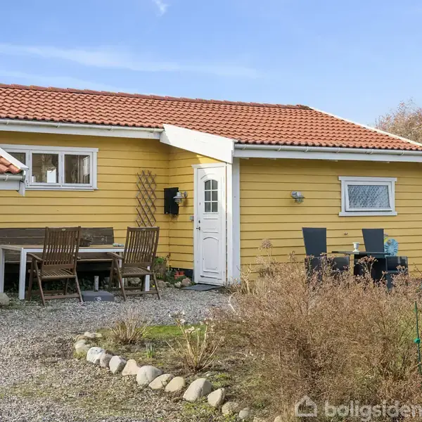 Et gult hus med rødt tag har en havedør og vindue. Foran huset står havemøbler på grus, omkranset af grøn beplantning og tørre buske under en klar blå himmel.