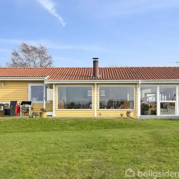 Et gult etplans hus står på en grøn græsplæne under en blå himmel. Der er store vinduer, en terrasse og en skorsten. Træer omkranser huset i baggrunden.