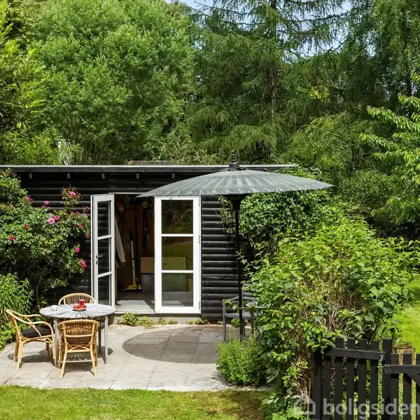Træhus med åben dør står i frodig have. Rundt cafébord med to stole og en parasol placeret på flisebelagt terrasse omkranset af grønne buske og træer.