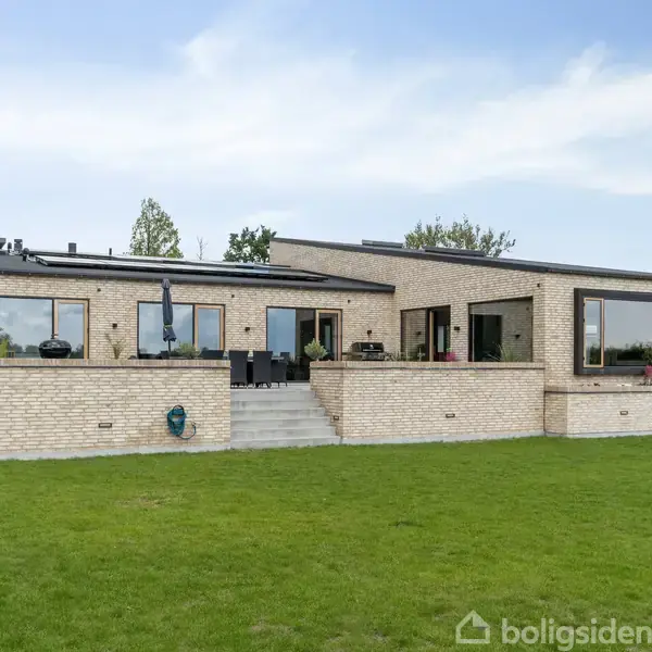 Hus i moderne stil med store vinduer står på en velplejet græsplæne. Der er en bred terrasse i sten med trapper. Omkring huset er der hække og få træer i baggrunden.