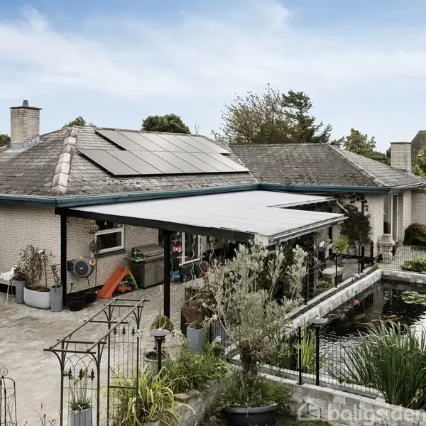 Et hus med solpaneler på taget står i en baggård, omgivet af planter og en stor fiskedam. En overdækket terrasse er placeret ved siden af dammen.