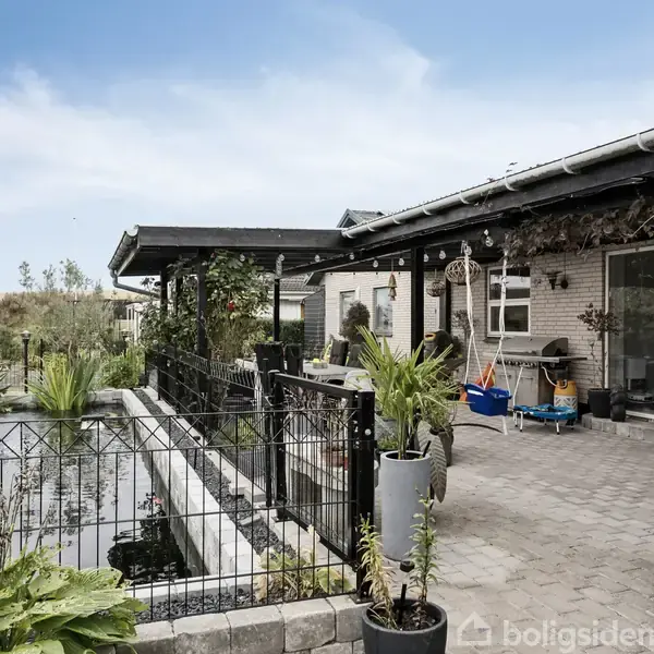 Hus omgivet af terrasse, hvor potteplanter står i ro. På venstre side ses en dam med metalgitter omkring. Himlen er blå med få skyer.