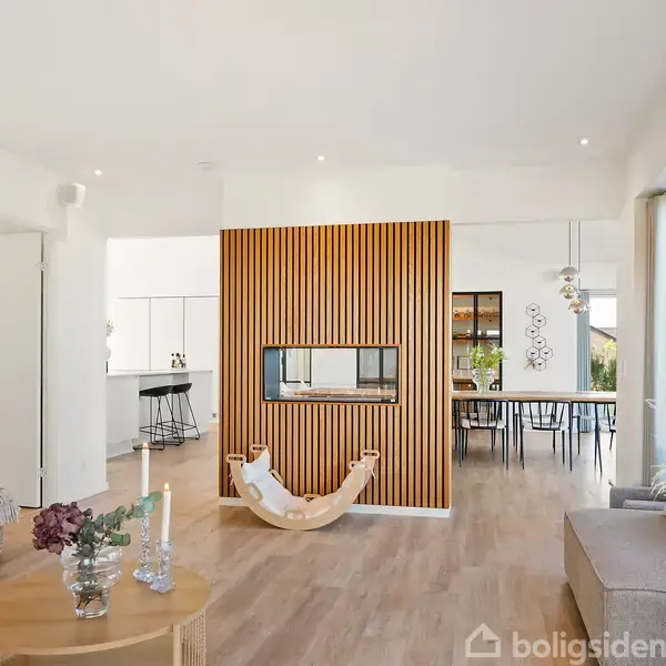 Stue med moderne indretning; åben pejs adskiller rummet, omgivet af sofaer og dekorative elementer. Køkken og spiseområde i baggrunden med store vinduer, der giver naturligt lys.