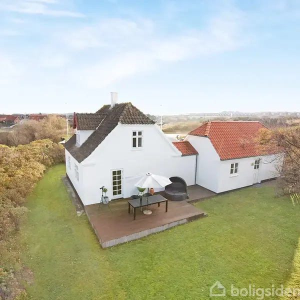 Hvidmalet hus med sort tag står på en grøn græsplæne med en terrasse, hvor et bord med parasol er placeret. Omgivet af buskads og med en klar himmel.