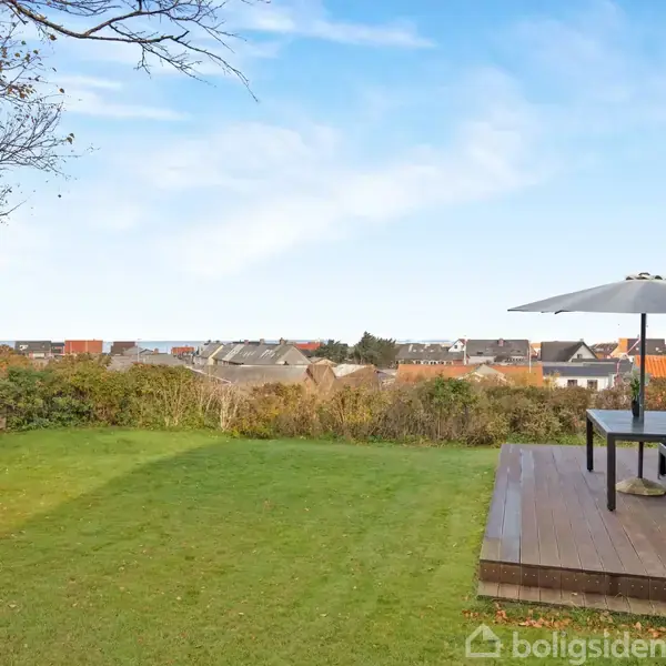 Terrasse med bord og parasol står på en træplatform. Grøn græsplæne strækker sig frem til en hæk, der adskiller haven fra et bymiljø med huse i baggrunden under en blå himmel.