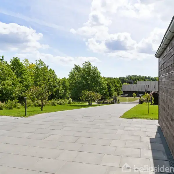 Flisebelagt gårdsplads strækker sig mod en have med træer, adjaceret til en moderne bygning i træ. Himlen er let overskyet med blå skyer.