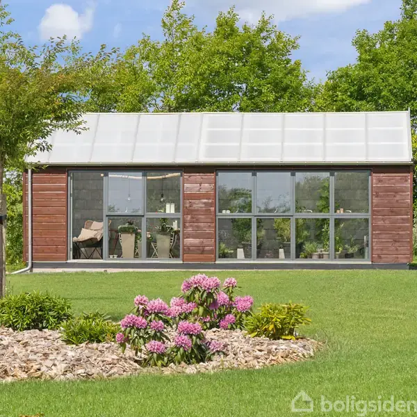 Et lille, moderne træhus med store glasdøre står på en velplejet græsplæne. Farverige blomster og grønne træer omgiver huset, der ses under en lyseblå himmel.