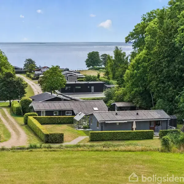 Sommerhuse omgivet af velholdte hække ligger langs en grusvej i et grønt landskab, med havet i baggrunden under en klar blå himmel.