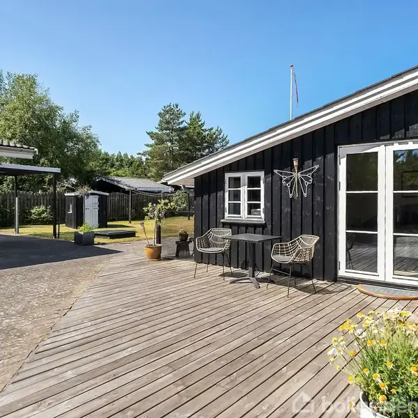 Sortmalet træhus med franske døre står på en træterrasse, omgivet af grønne planter og udendørs møbler. Haven inkluderes i en solrig, fredfyldt atmosfære.