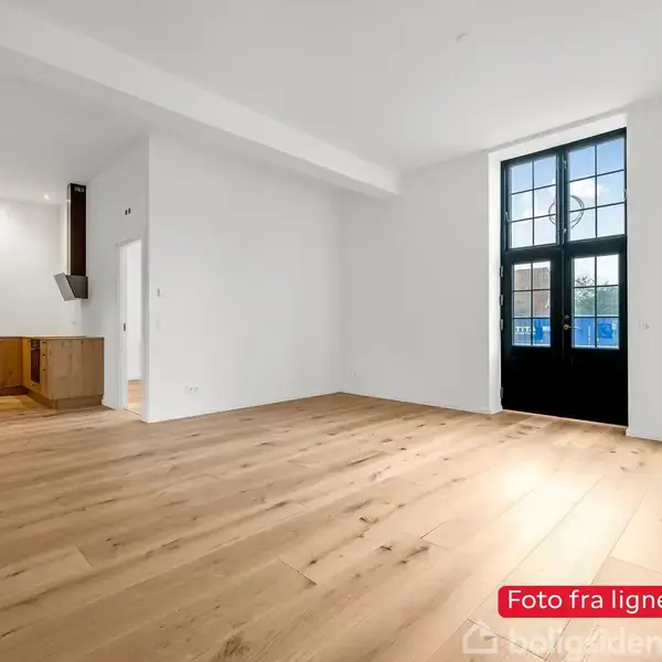 Lyst trægulv breder sig i en stue med hvide vægge. Store glasdøre fører ud til en balkon. Tekst siger: "Foto fra lignende bolig". Venstre side har et åbent køkken med træskabe.