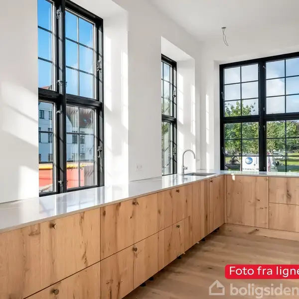 Køkkenbordet har en træoverflade med skuffer, placeret under store rektangulære vinduer, der tillader naturligt lys at strømme ind. Tekst: "Foto fra lignende bolig."