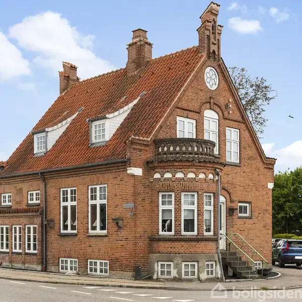 Rødt murstenshus med trekantet gavl og udsmykning foran. En trappe fører op til en indgang. En blå bil er parkeret ved siden af huset. Skilt: "Din Lokale Sagfører Over 100 År".