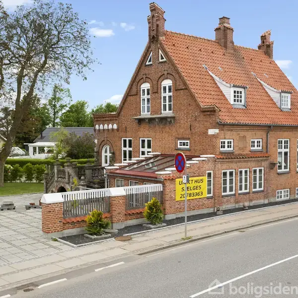 Rødt murstenshus med spidst tag sælges. Beliggende ved en vej omgivet af få træer og græs. Skiltet foran huset siger: "L. Haus til salg 20 78 36 33."
