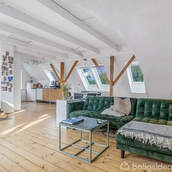 En grøn sofa står i et lyst loftsrum med trægulv. Der er skråvinduer, synlige træbjælker, og et lille køkkenområde i baggrunden. En brændeovn ses til venstre.