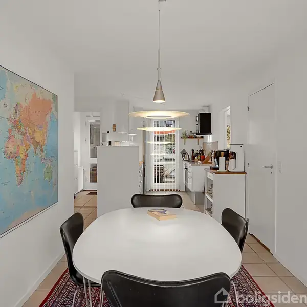 Ovalt bord omgivet af stole står i et lyst køkken. Verdenskort hænger på væggen til venstre. Lampe hænger over bordet, og en dør fører til andre rum.