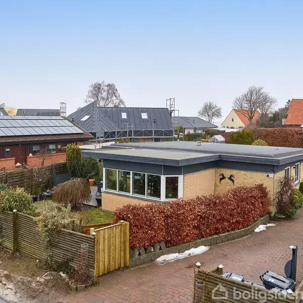 En moderne, lavtliggende bungalow med fladt tag er omgivet af en have med hække og hegn i forgrunden. Naboens hus har solpaneler, og et nyt hus er under opførelse i baggrunden.