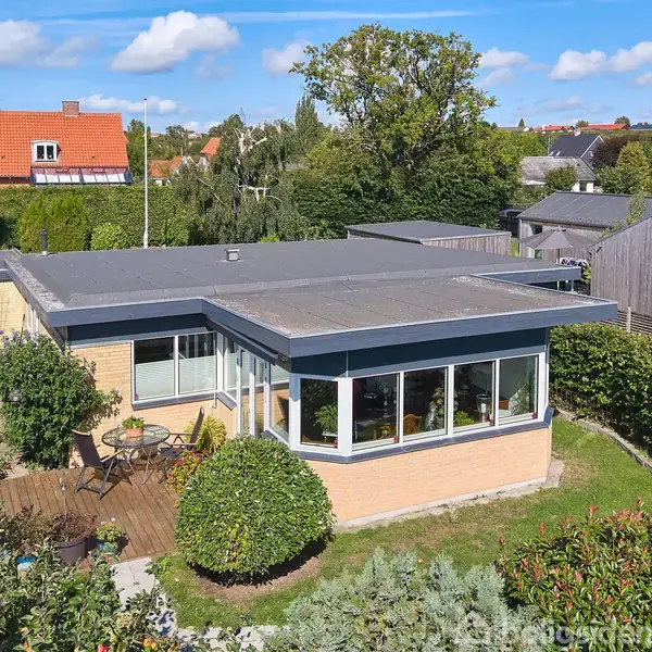 Et étplanshus med fladt tag står midt i en velplejet have med græsplæne og buske. Der er en terrasse i træ med havemøbler. Baggrunden viser flere nabotage.
