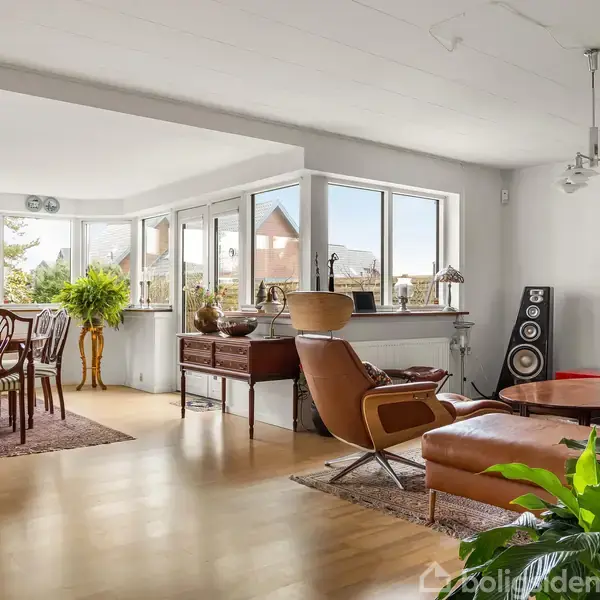 En lys stue med lænestol og sofabord står i forgrunden. Et spisebord med stole er placeret ved store vinduer. Grønne planter pynter rummet, der har trægulve og moderne indretning.