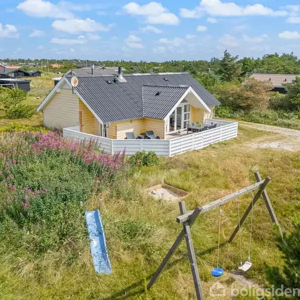 Et gult træhus med mørkt tag står i en grønt område. En terrasse er omgivet af et hvidt hegn. En gynge og rutsjebane ses i haven.