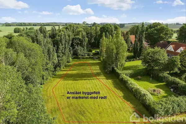 Græsareal markeret med rød linje indikerer byggefelt. Omkranset af træer og buske i landlig område med huse i baggrunden. Tekst: "Anslået byggefelt er markeret med rødt".