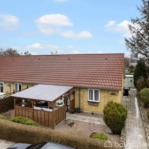 En bungalow med teglstenstag og carport står i en velholdt have omkranset af hække. Solen skinner, og en sti fører op til hoveddøren. Husnummer 287 er synligt.