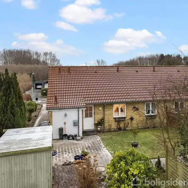 En gul murstensbygning med rødt tegltag står ved siden af en lille, velplejet have. Foran er et skur og flisebelagt sti. Bagved ses en grusvej omkranset af buske.