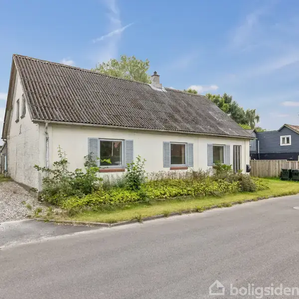Et hvidt hus med skråt tag og to vinduer vender ud mod en stille vej. Omkranset af græs og buske i et villakvarter.