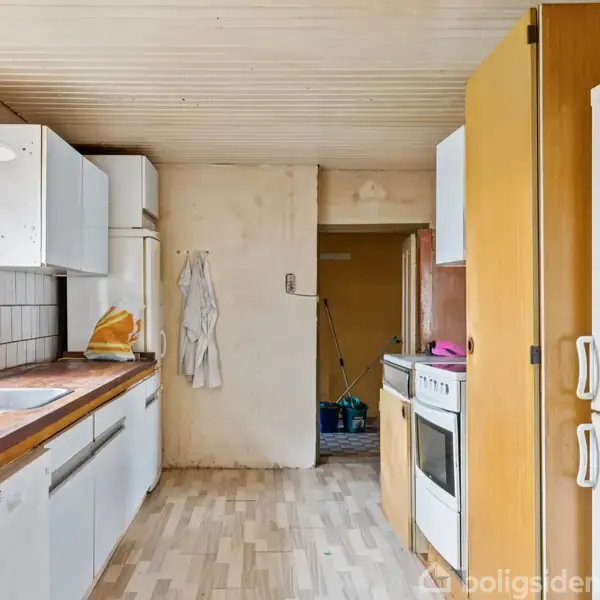 Et lyst køkken med et stort vindue til venstre, indeholder en vask og køleskab. Et komfur med ovn står til højre, omgivet af skabe. Ingen tekst til stede.