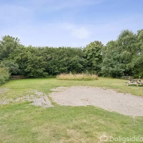En græsplæne med en grusplads i midten, omgivet af buske og træer, og en picnicbænk står til højre, under en blå himmel.