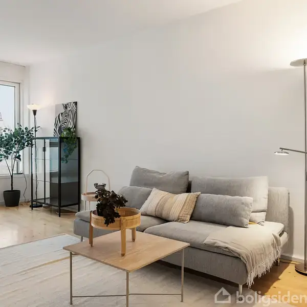 Grå sofa med puder ved gulvlampe i moderne stue; lys træstue med stort vindue. Spisebord og stole i baggrunden. Grøn plante tæt på glasvitrine.