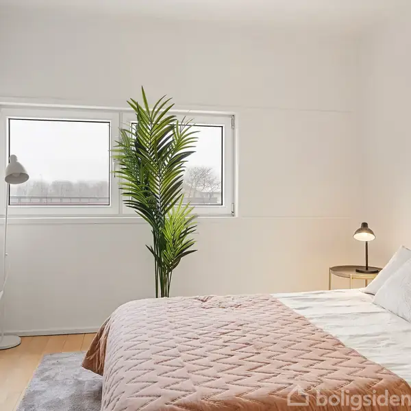 Soveværelse med en grå dobbeltseng dækket af puder og et quiltet sengetæppe. Til venstre ses et højt spejl, en gulvlampe og en plante under to vinduer.