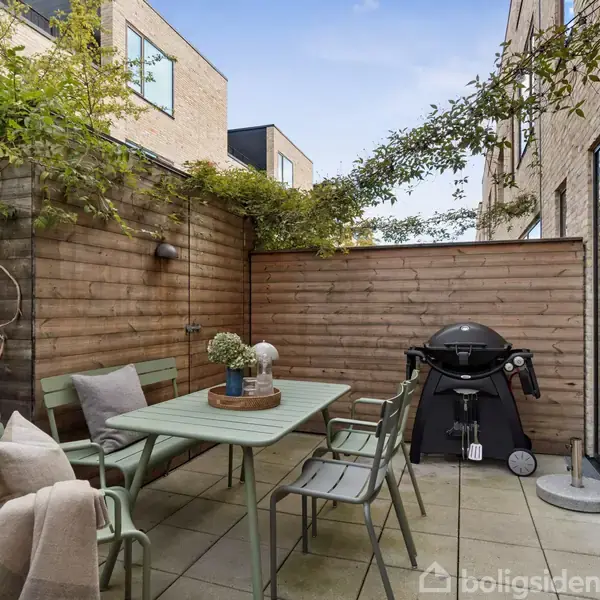 Terrasse med spisebord og stole i grøn metal. En gasgrill står ved siden af den lukkede træplanke-skovl. Planter klatrer op ad væggene, omgivet af moderne hjem.