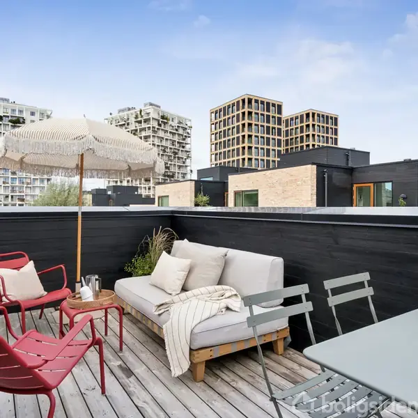 Terrasse med sofa og røde stole placeret omkring et bord med paraply, omgivet af moderne bygninger i baggrunden. En grøn plante og tæppe skaber en hyggelig atmosfære.
