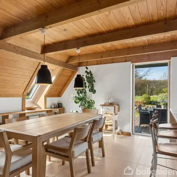 Træspisebord med stole står i et lyst rum. Bordet har to hængelamper over sig. Store skråvinduer og en åben dør fører ud til en terrasse med planteudsigt.