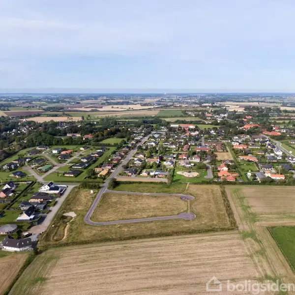En landsby set oppefra med spredte huse, grønne områder og omkringliggende marker. Vejnettet skaber en struktureret opdeling af boligerne i et ellers landligt landskab.