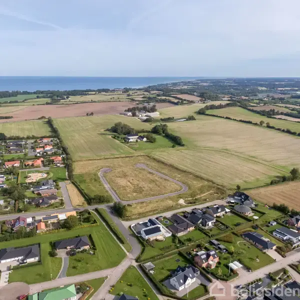 Landskab med marker og huse spredt i forgrunden, beliggende tæt på en kystlinje. En vej omgiver et udstykket område i midten, hvilket indikerer planlagt byggeri.