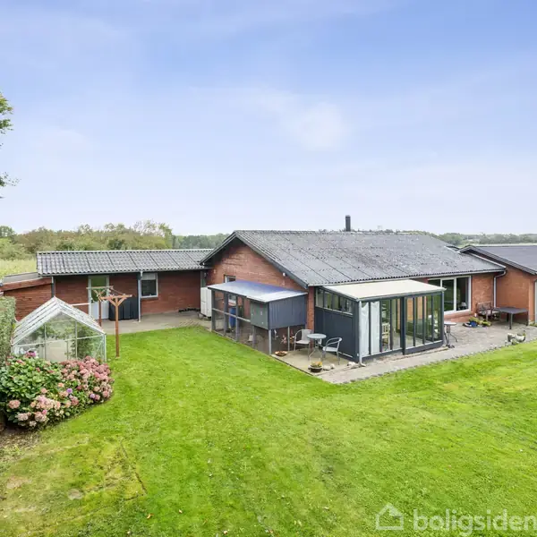 Hus med røde mursten og sort tag omgivet af stor græsplæne; ved siden er et drivhus. I baggrunden ses åbne marker og træer under en blå himmel.