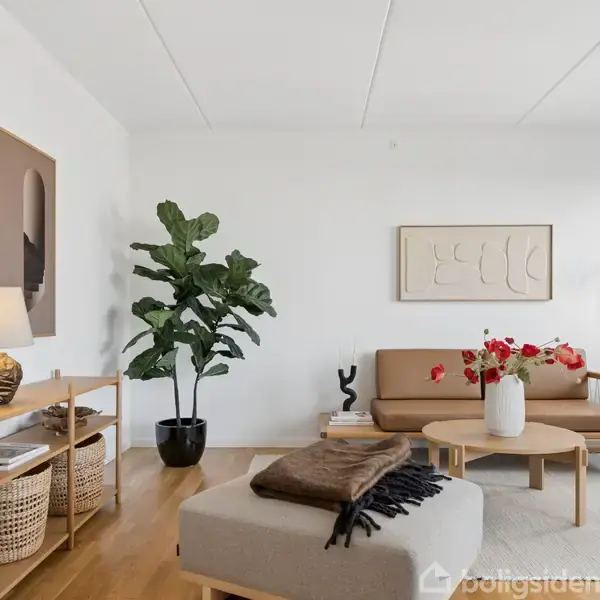 Stue med sofa, sofabord og stor plante. Sofaen er brun med puder, bordet har en vase med blomster. Hylde med lamper og kurve står ved siden af.