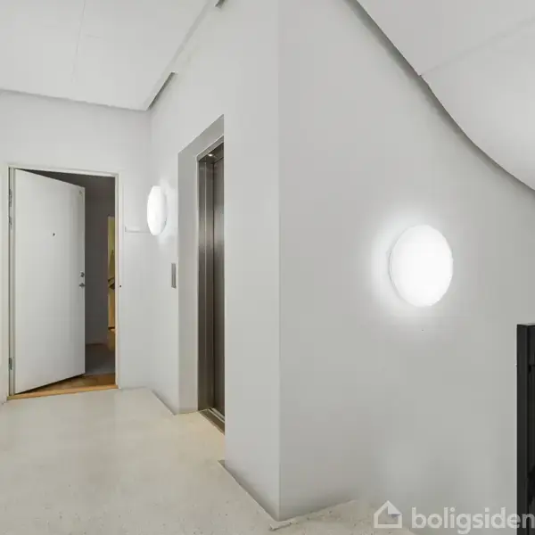 Elevator står åben i en lys opgang med hvide vægge. To runde væglamper oplyser etagen. En hvid dør er åben til højre i baggrunden.