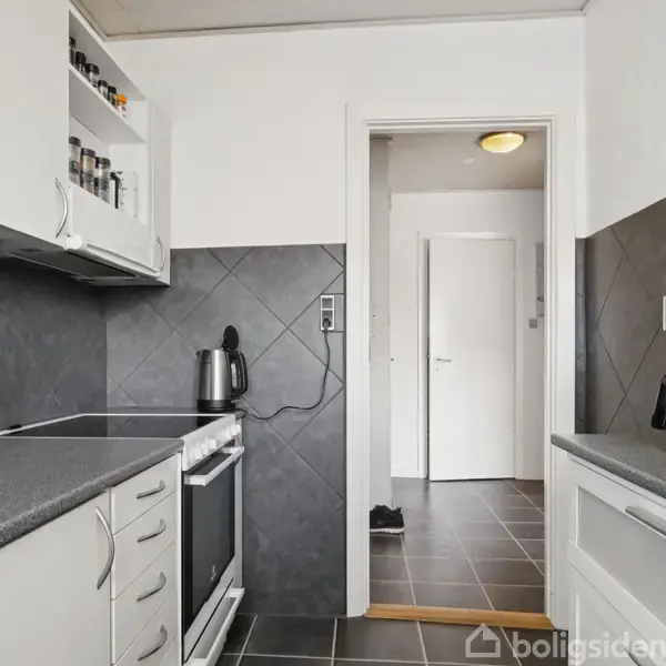 Et køkken med hvide skabe og sorte fliser. En elkedel står på køkkenbordet ved siden af en ovn. En kaffemaskine ses til højre. En dør fører til en lys gang.