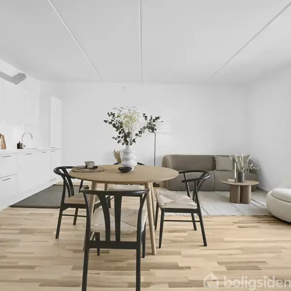Et lyst køkken-alrum med trægulv, hvidt køkken med indbygningsovn, rundt spisebord med fire stole og en vase med blomster. Stueområde med beige sofa, gulvlampe og lænestol.