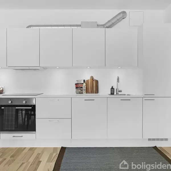 Hvidt køkken med skabe, indbygningsovn og komfur. Køkkenredskaber er placeret på bordpladen. Trægulv og tæppe foran køkkenet. Ventilationsrør ses øverst.