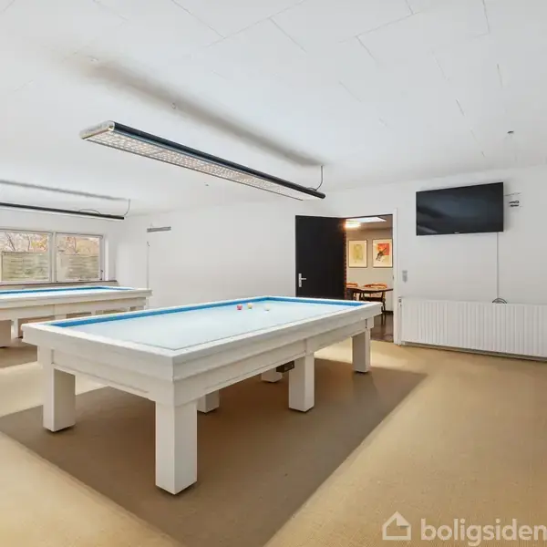 Poolborde står opstillet i et lyst rum med beige gulv og hvide vægge. Der er en vægmonteret tv-skærm og en række sorte stole langs væggen.