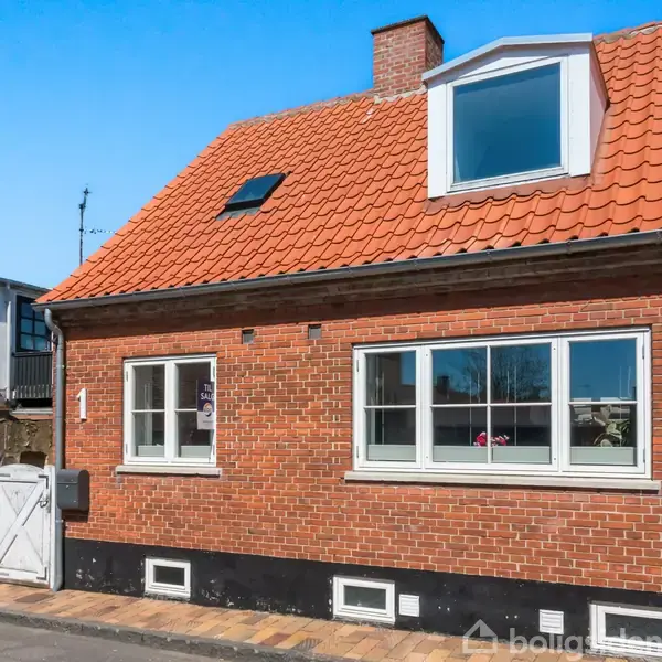 Rødt murstenshus med hvid rammevinduer og skråt tag med en kvist. Dobbeltfacade og hvid trælåge i forgrunden. Klar blå himmel i baggrunden. Nummer "1" er på facaden.
