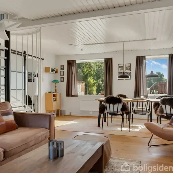 Stue med lædersofa og lænestol, sofabord i træ, spiraltrappe til venstre, spisebord med stole i baggrunden, store vinduer med udsigt til have og grønne omgivelser.