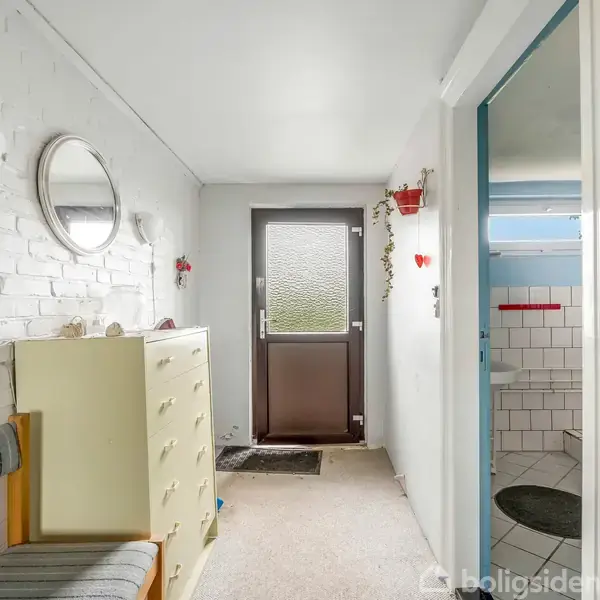 Entre med hvidmalede murstensvægge; en kommode står langs væggen under et rundt spejl. Dør i enden leder udenfor. Et åbent badeværelse er til højre med blå fliser.