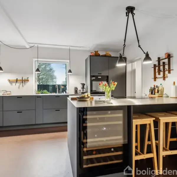 Et moderne køkken med grå skabe og en stor køkkenø med indbygget vinkøleskab. Tre træstole står ved øen. Blomster og belysning skaber en hyggelig atmosfære.