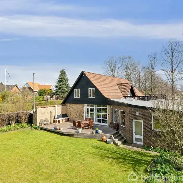 Et murstenshus med rødt tegltag står stille i en frodig have. Der er en stor terrasse med møbler omkring, omgivet af træer og buske under en klar blå himmel.