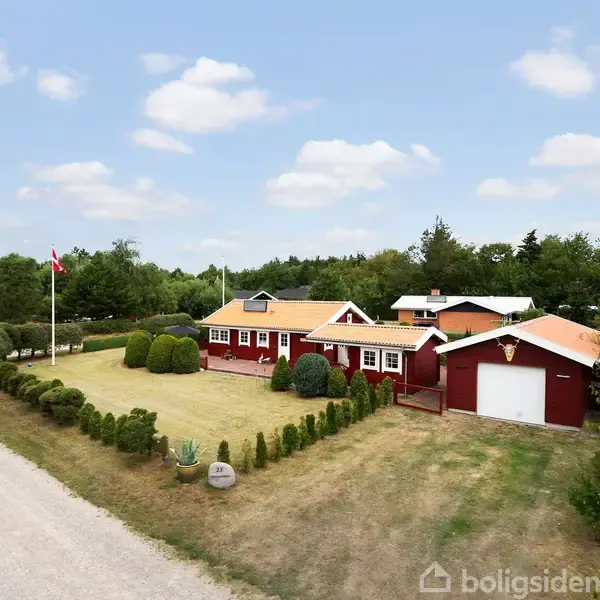 Rødt hus med hvide vindueskarme står stille i en have med velholdt buskads. En flagstang med flag er på græsplænen. Grusvej fører forbi i et grønt landskab.