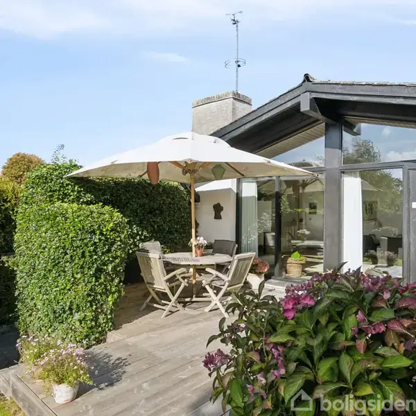 En terrasse med en rund bord og fire stole under en stor parasol. Omkring er der grønne buske og planter, og huset har store glaspartier.